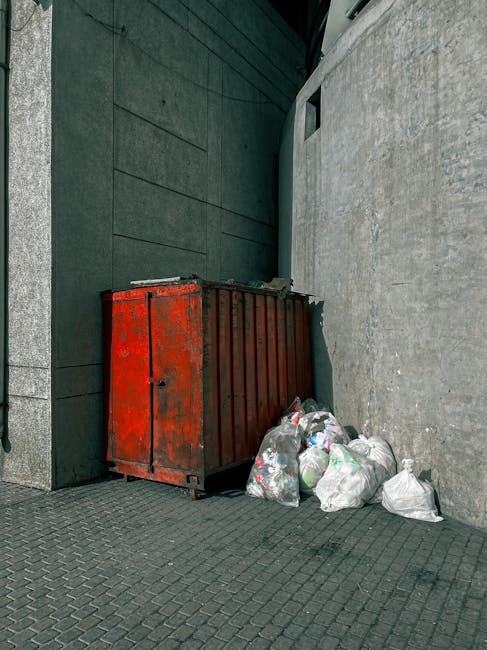 A weathered red metal dumpster with visible rust and peeling paint is positioned against a dark, modern building wall with horizontal paneling on the left. The dumpster's lid is slightly bent and part