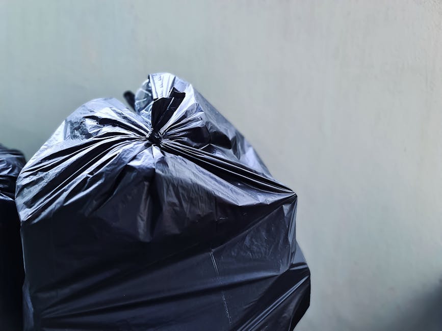 A weathered red metal dumpster with visible rust and peeling paint is positioned against a dark, modern building wall with horizontal paneling on the left. The dumpster's lid is slightly bent and part