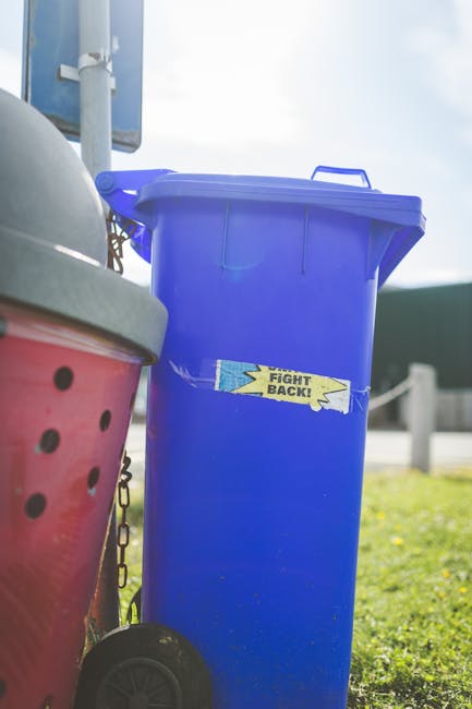 In the image, a large blue wheelie bin with a closed lid is positioned outdoors on a grassy area, with a small handle visible on the top edge of the lid. The bin is made of plastic with a smooth, slig